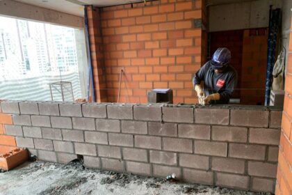 Operário assentando blocos estruturais de concreto em obra residencial, com paredes de tijolos ao fundo, demonstrando técnicas modernas de alvenaria.