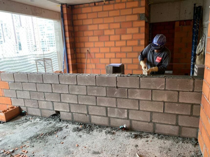 Operário assentando blocos estruturais de concreto em obra residencial, com paredes de tijolos ao fundo, demonstrando técnicas modernas de alvenaria.