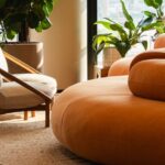 Ambiente corporativo moderno com sofá arredondado laranja, poltrona bege, plantas naturais e luz natural entrando por grandes janelas, destacando o conceito de biofilia nos escritórios.