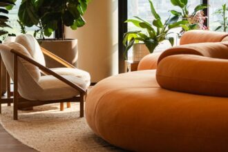 Ambiente corporativo moderno com sofá arredondado laranja, poltrona bege, plantas naturais e luz natural entrando por grandes janelas, destacando o conceito de biofilia nos escritórios.
