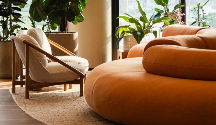 Ambiente corporativo moderno com sofá arredondado laranja, poltrona bege, plantas naturais e luz natural entrando por grandes janelas, destacando o conceito de biofilia nos escritórios.