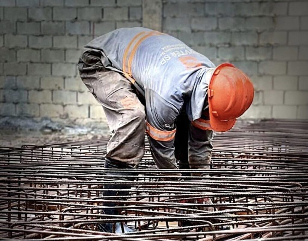 Profissional da construção civil com capacete laranja trabalha na montagem de armação de aço para fundação, demonstrando precisão e atenção aos detalhes.