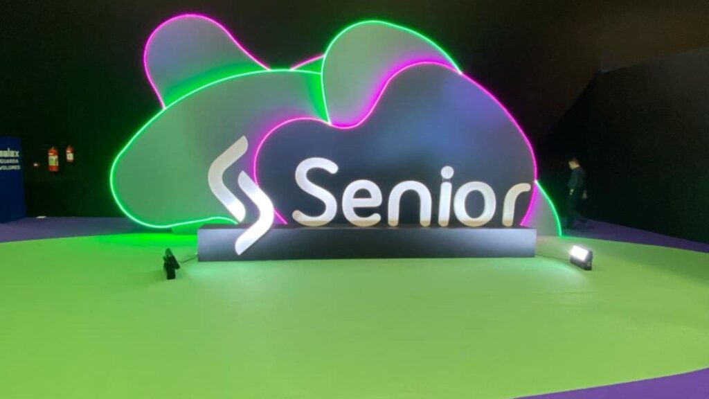 Escultura iluminada com o logo da Senior Sistemas em destaque no espaço do Senior Experience 2026, sobre piso verde e roxo, simbolizando inovação e identidade visual da marca.