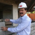 Milton Bigucci, presidente da MBigucci, sorrindo enquanto assenta bloco de concreto em obra, usando capacete de segurança branco com logotipo da empresa. Foto de 2016, crédito Celso Lima.