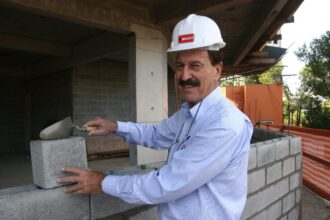 Milton Bigucci, presidente da MBigucci, sorrindo enquanto assenta bloco de concreto em obra, usando capacete de segurança branco com logotipo da empresa. Foto de 2016, crédito Celso Lima.