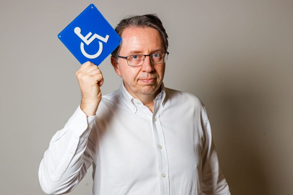 Homem de óculos e camisa branca segura placa azul de acessibilidade com expressão séria, destacando a importância da inclusão na construção civil.