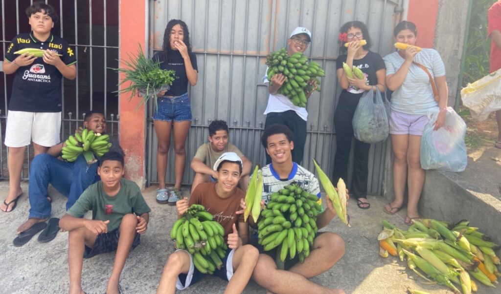 Grupo de crianças e adolescentes sorrindo, segurando cachos de bananas, espigas de milho e hortaliças frescas em frente a um portão, representando colheita comunitária e integração social.