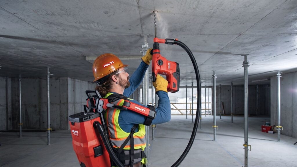 Profissional da construção civil utiliza martelete Hilti com sistema de extração de pó para perfuração no teto, equipado com EPI completo em ambiente de obra. Imagem Ilustrativa criada pela Equipe C3.
