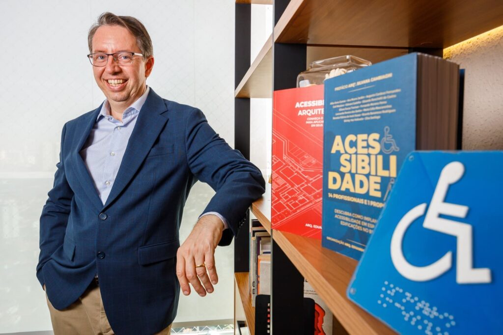 Homem de terno azul sorri ao lado de estante com livros sobre acessibilidade e placa azul de acessibilidade em destaque, representando liderança e conhecimento em inclusão na construção civil.