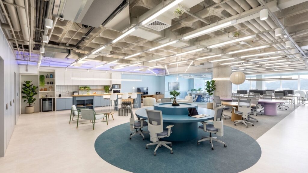 Ambiente corporativo moderno da Space, com layout aberto, mesas redondas colaborativas, cadeiras ergonômicas, iluminação natural e artificial, plantas naturais e área de convivência integrada à copa, promovendo bem-estar, inovação e produtividade.