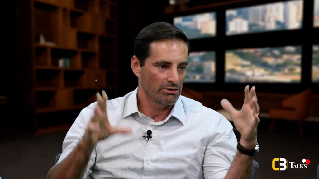 Rodrigo Rocha, Head de Empreendimentos Residenciais da Rocontec, gesticula com as mãos enquanto fala durante sua participação no C3 Talks, em ambiente corporativo moderno com painéis de cidade ao fundo.