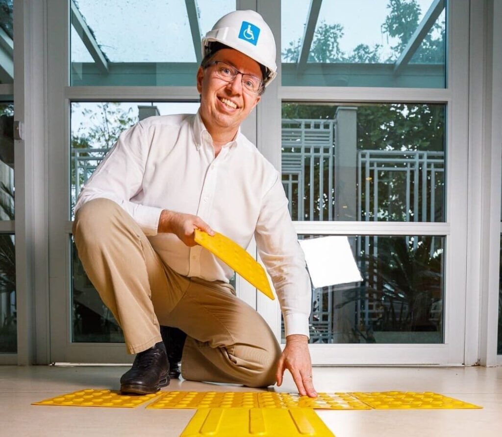 Homem de capacete branco com símbolo de acessibilidade instala piso tátil amarelo em ambiente interno, promovendo inclusão na construção civil.