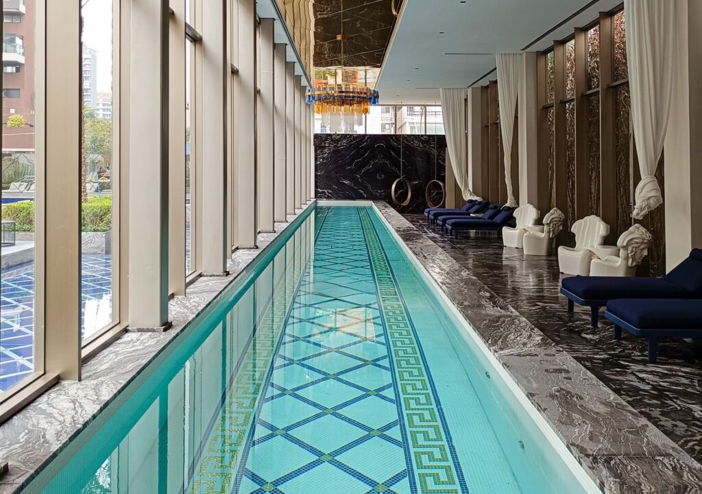 Piscina interna de alto padrão no Villa Residence Lavvi Praça Central, com revestimento em pedras naturais, espreguiçadeiras sofisticadas e grandes janelas que proporcionam iluminação natural e vista para o paisagismo externo.