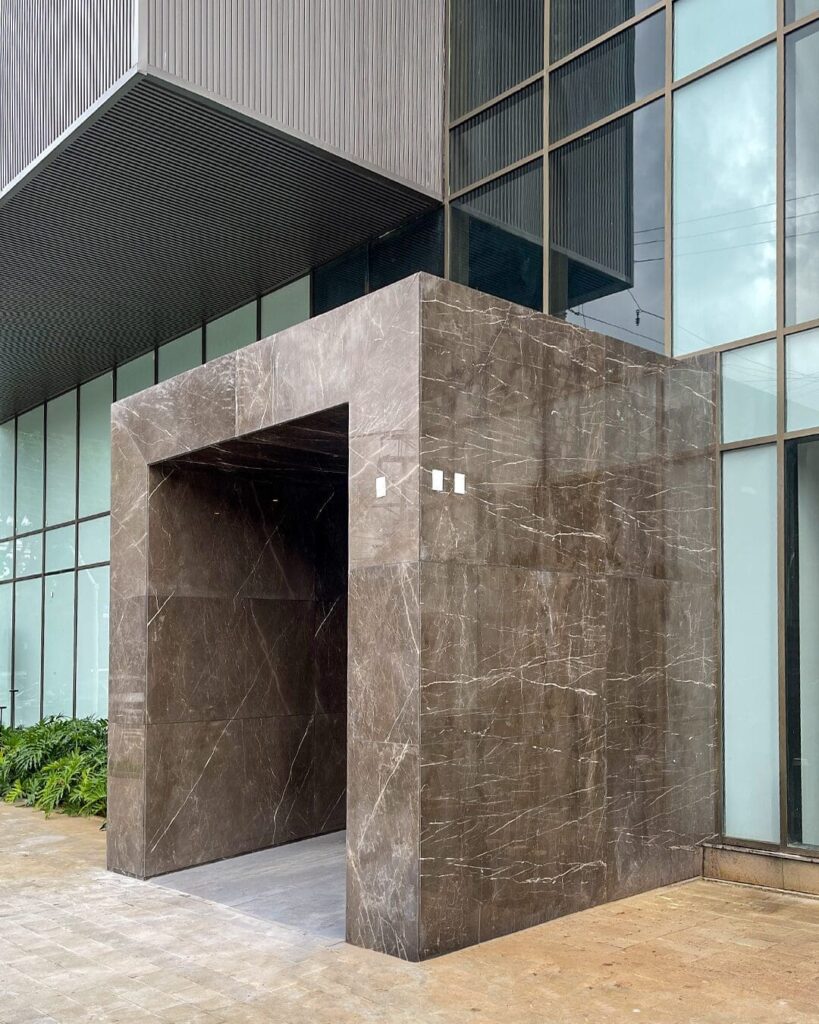 Entrada do edifício Park Avenue revestida com grandes placas de pedra natural escura, destacando linhas retas, acabamento sofisticado e arquitetura contemporânea.
