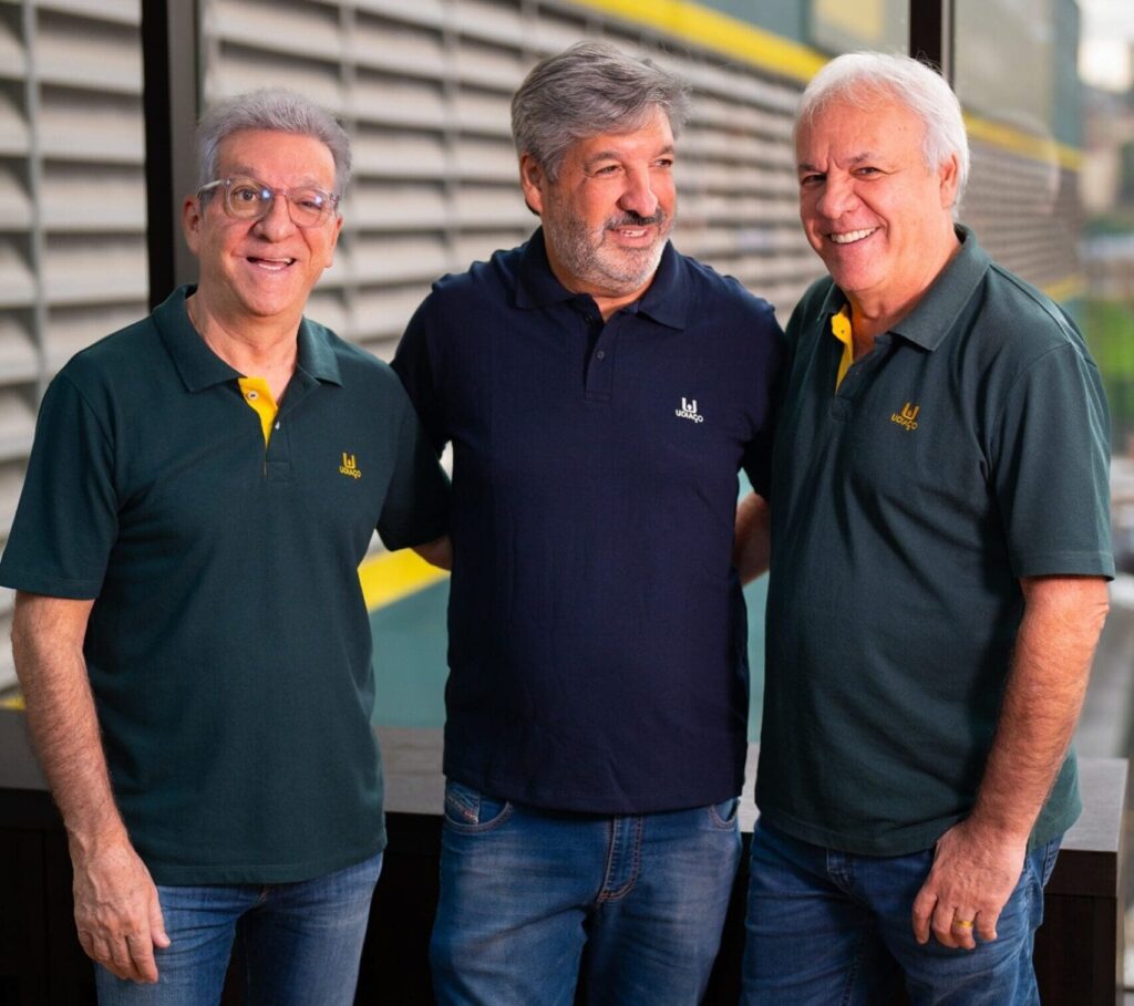 Três fundadores da Udiaço, sorridentes e de braços dados, posam juntos em ambiente interno com fachada industrial da empresa ao fundo, simbolizando união, liderança e tradição no setor de aço para construção civil.