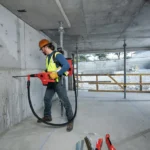 Trabalhador da construção civil utiliza martelete Hilti com sistema de extração de pó em obra, vestindo EPI completo.