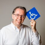 Homem sorridente de óculos e camisa branca segura placa azul de acessibilidade, simbolizando inclusão na construção civil.