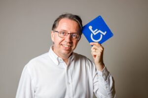 Homem sorridente de óculos e camisa branca segura placa azul de acessibilidade, simbolizando inclusão na construção civil.