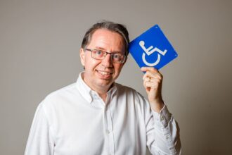 Homem sorridente de óculos e camisa branca segura placa azul de acessibilidade, simbolizando inclusão na construção civil.