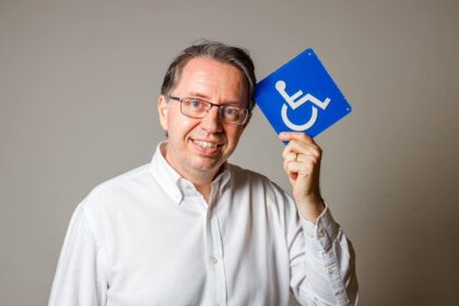 Homem sorridente de óculos e camisa branca segura placa azul de acessibilidade, simbolizando inclusão na construção civil.