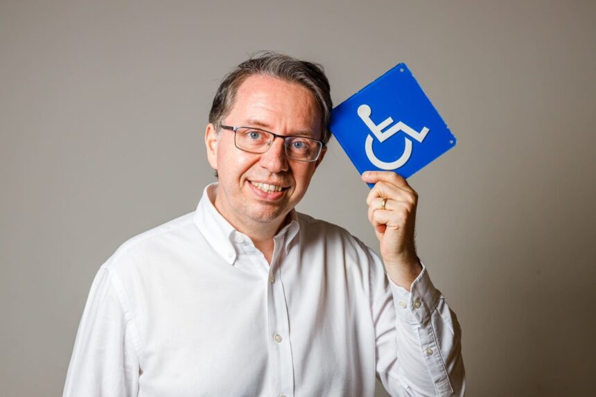 Homem sorridente de óculos e camisa branca segura placa azul de acessibilidade, simbolizando inclusão na construção civil.