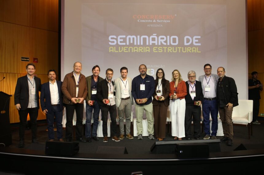 Grupo de palestrantes e organizadores do Seminário de Alvenaria Estrutural do C3 posam juntos no palco do auditório, com o telão ao fundo exibindo o nome do evento.