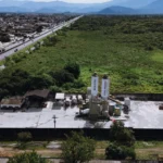 Vista aérea da filial Guarujá da Concreserv, mostrando a unidade de concreto usinado ao lado de uma rodovia movimentada e áreas verdes, com a cidade e montanhas ao fundo.