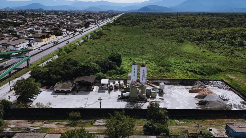 Vista aérea da filial Guarujá da Concreserv, mostrando a unidade de concreto usinado ao lado de uma rodovia movimentada e áreas verdes, com a cidade e montanhas ao fundo.