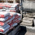 Sacos de argamassa estrutural AAE8 empilhados em paletes em um canteiro de obras, com paredes de blocos em construção ao fundo.