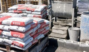 Sacos de argamassa estrutural AAE8 empilhados em paletes em um canteiro de obras, com paredes de blocos em construção ao fundo.