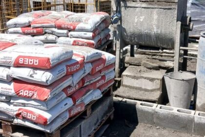 Sacos de argamassa estrutural AAE8 empilhados em paletes em um canteiro de obras, com paredes de blocos em construção ao fundo.