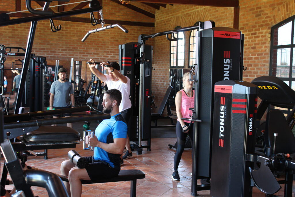 Pessoas treinando em uma academia equipada com aparelhos modernos de musculação, ambiente com paredes de tijolo aparente e iluminação natural.