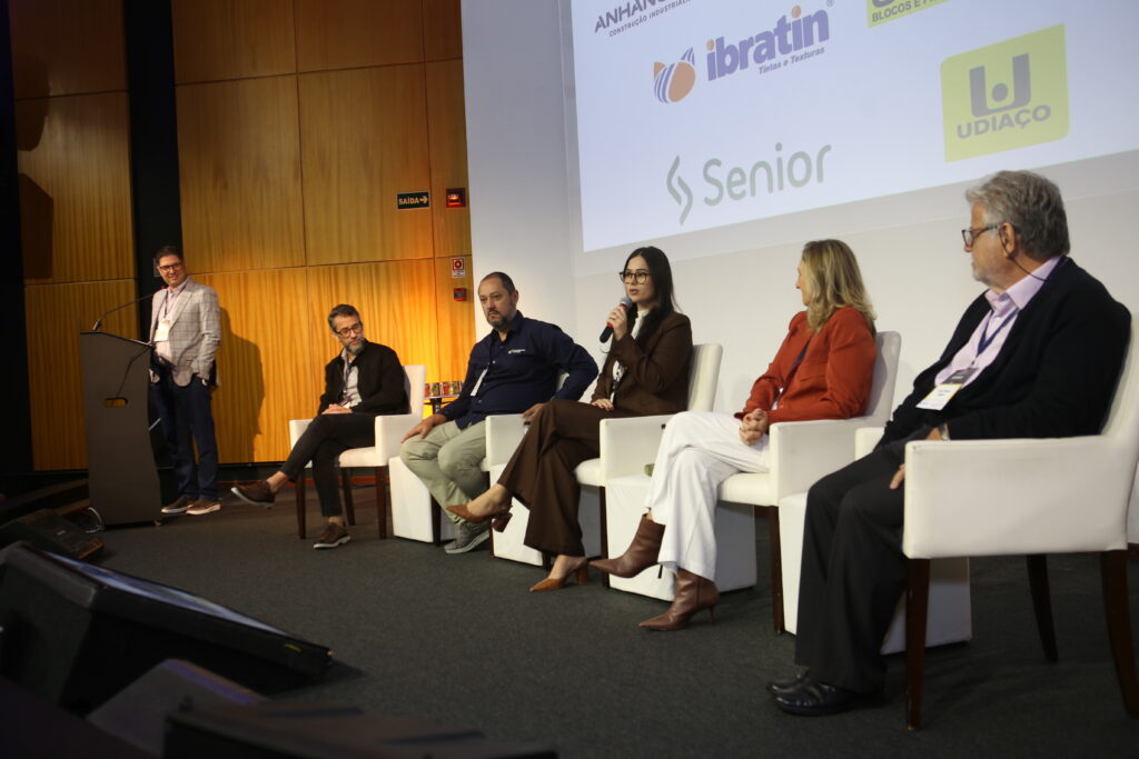 Painelistas e mediador debatem industrialização da alvenaria estrutural no palco do Seminário do C3, diante de um auditório lotado de profissionais da construção civil, com destaque para a interação e diversidade dos participantes.