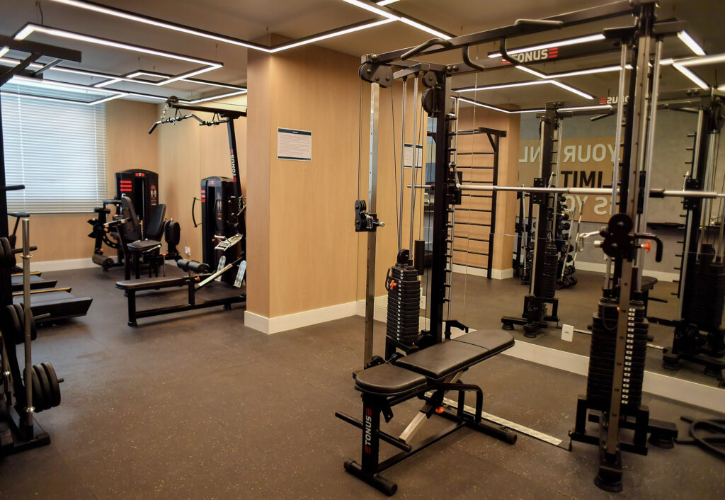 Academia compacta e moderna com equipamentos de musculação, espelhos amplos, iluminação em LED e acabamento em madeira clara, ideal para empreendimentos residenciais.