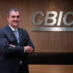 Homem de terno escuro, sorrindo, posando de braços cruzados em frente ao logo da CBIC em uma parede de madeira.