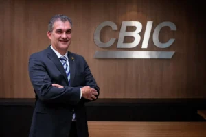 Homem de terno escuro, sorrindo, posando de braços cruzados em frente ao logo da CBIC em uma parede de madeira.