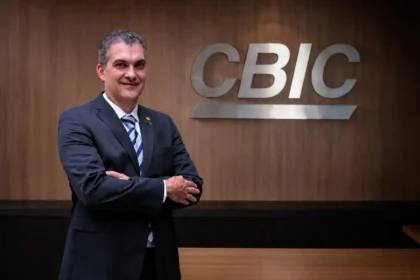 Homem de terno escuro, sorrindo, posando de braços cruzados em frente ao logo da CBIC em uma parede de madeira.