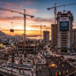 Canteiro de obras moderno com trabalhadores, gruas e prédios em construção, destacando as logomarcas da CBIC e do C3, ao entardecer, simbolizando inovação, qualificação e desafios da construção civil brasileira.