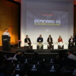 Auditório lotado no Milenium Centro de Convenções durante o Painel 2 do Seminário de Alvenaria Estrutural do C3, com profissionais da construção civil atentos ao debate sobre industrialização da alvenaria estrutural, painelistas no palco e logomarcas de patrocinadores em destaque.