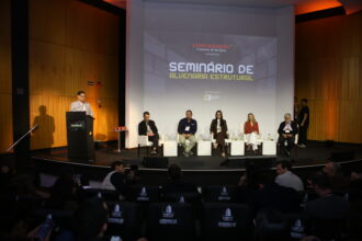 Auditório lotado no Milenium Centro de Convenções durante o Painel 2 do Seminário de Alvenaria Estrutural do C3, com profissionais da construção civil atentos ao debate sobre industrialização da alvenaria estrutural, painelistas no palco e logomarcas de patrocinadores em destaque.