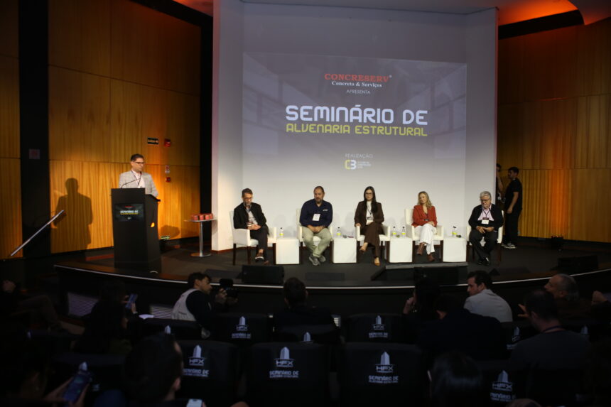 Auditório lotado no Milenium Centro de Convenções durante o Painel 2 do Seminário de Alvenaria Estrutural do C3, com profissionais da construção civil atentos ao debate sobre industrialização da alvenaria estrutural, painelistas no palco e logomarcas de patrocinadores em destaque.